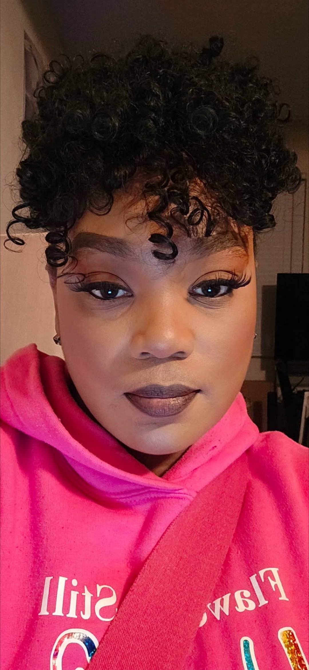 Close-up selfie of a woman wearing a bright pink hoodie with curly black hair styled in an updo, with defined eye makeup and brown lip color
